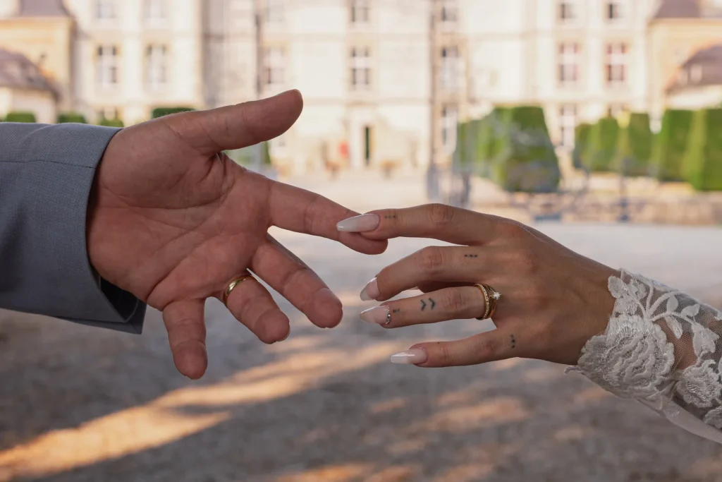 Gros plan sur les mains des mariés qui se rejoignent, alliances visibles, devant le château de Vitry-la-Ville, mariage dans la Marne